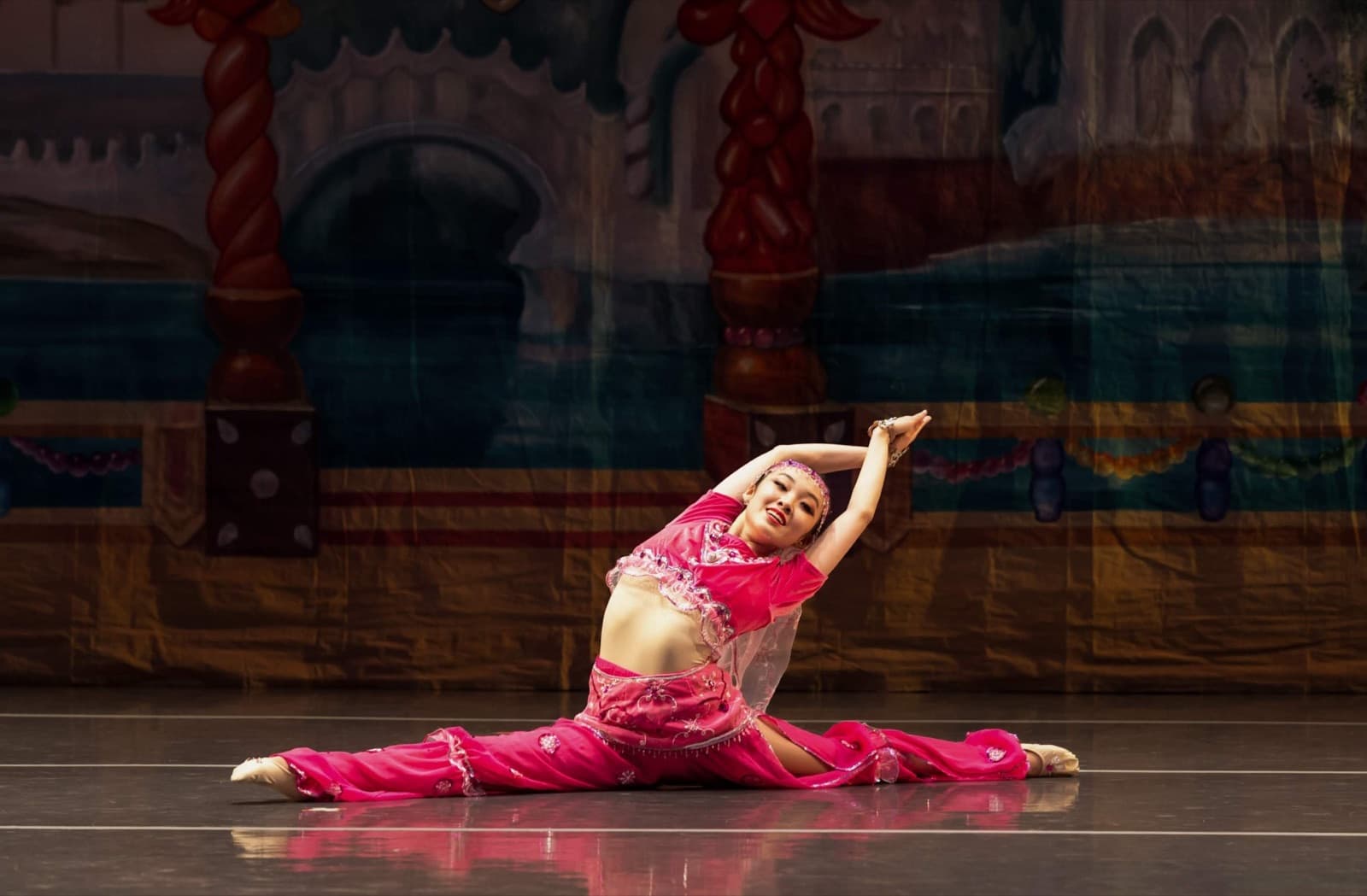 A dancer in a pink beaded costume sitting in a full split on stage, arms curved overhead, before a painted palace backdrop.