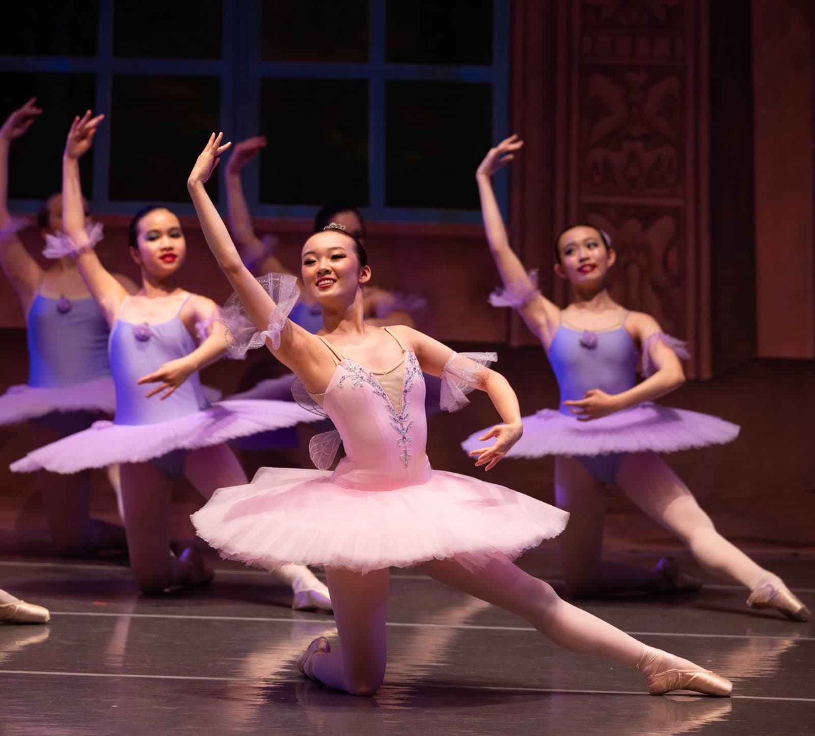 A ballerina in a pink tutu kneeling in a lunge with one arm raised, in front of a line of dancers in lilac tutus.