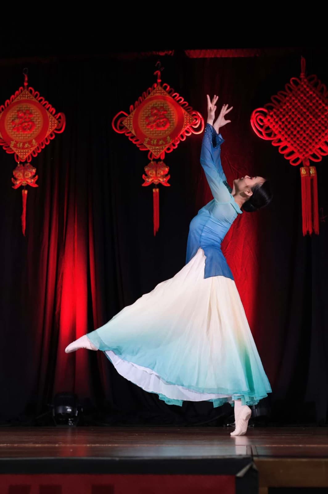 A dancer in a blue top and long white-to-teal skirt arching back with both arms reaching up, one leg extended behind, before red Chinese knot decorations on a dark curtain.