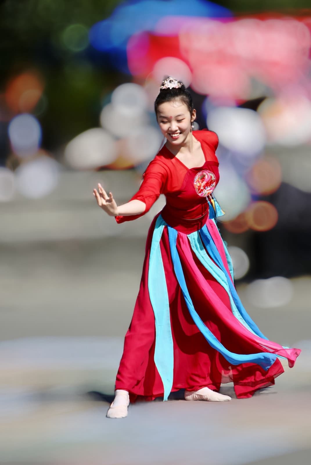 A smiling dancer outdoors in a red dress with streaming blue and pink ribbons, one hand extended, against a blurred colorful background.