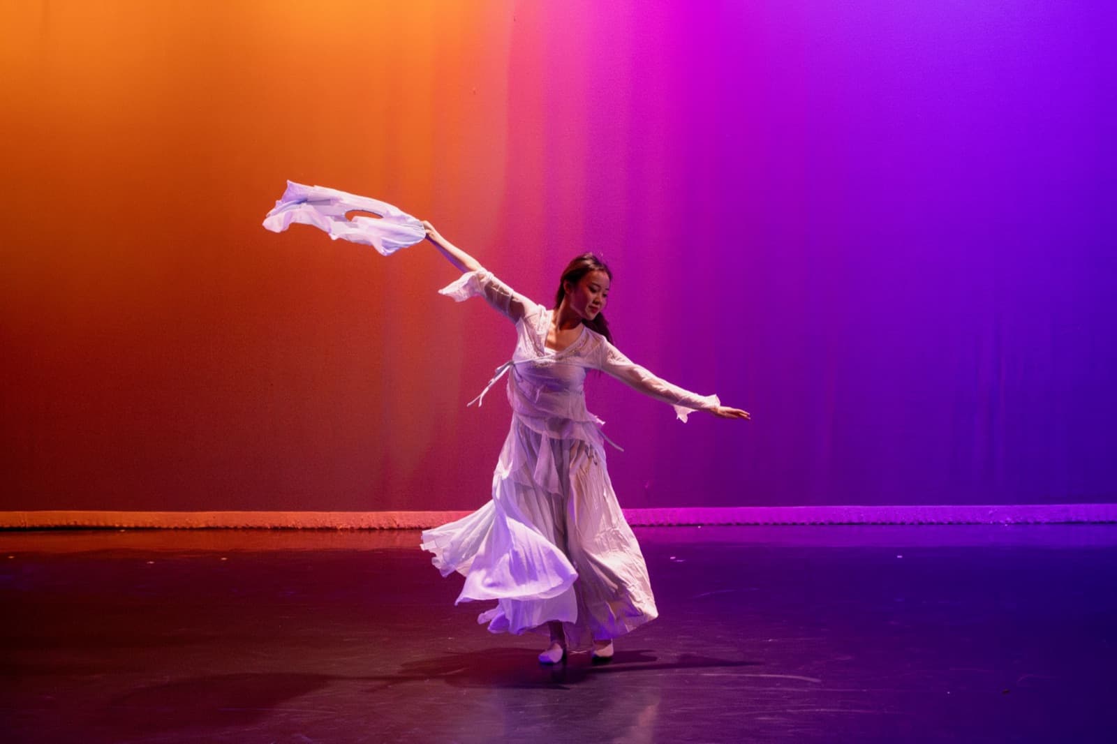 A dancer in flowing white turning across the stage, one arm sweeping a length of white silk overhead, against an orange-to-violet lit backdrop.