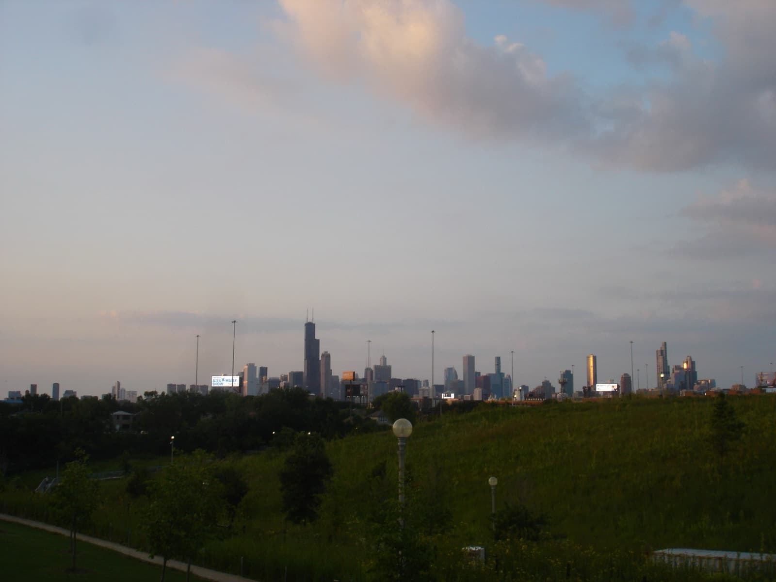 The Chicago skyline at dusk seen across a grassy hill and park lamps, under a wide pale sky with pink-lit clouds.